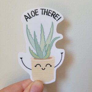May include: A white sticker with a cartoon aloe plant in a pot. The plant has green leaves and a smiling face. The text "Aloe There!" is written in black above the plant.