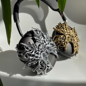 May include: A pair of black headphones with silver and gold decorative earcups. The earcups feature intricate, detailed designs.