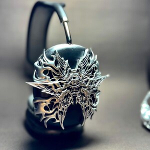 May include: Black headphones with a silver, ornate, butterfly-shaped design featuring a central eye motif.