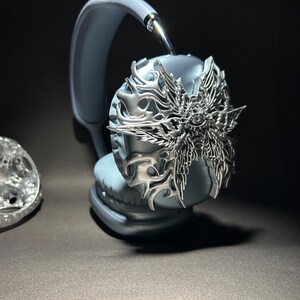 May include: A pair of gray headphones with a silver decorative cover featuring a winged, eye-like design.