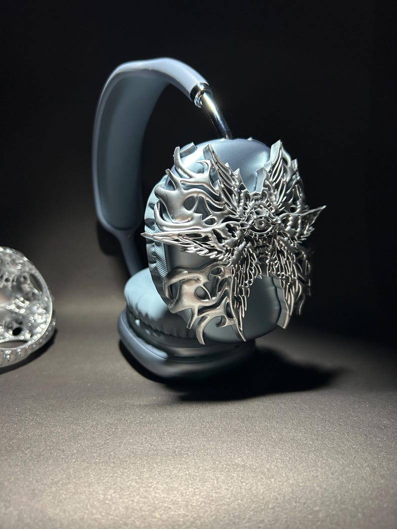 May include: A pair of gray headphones with a silver decorative cover featuring a winged, eye-like design.
