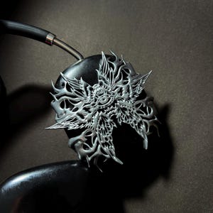 May include: Silver headphone earcup with a detailed, intricate, and ornate design of a stylized butterfly with a central eye.
