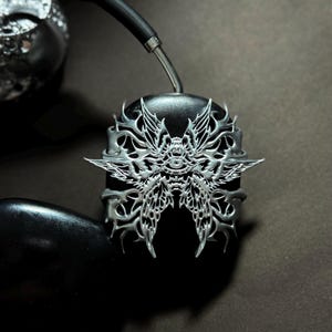May include: A pair of black headphones with a silver butterfly design on the earcup. The butterfly design is intricate and detailed, with a delicate, filigree-like appearance.
