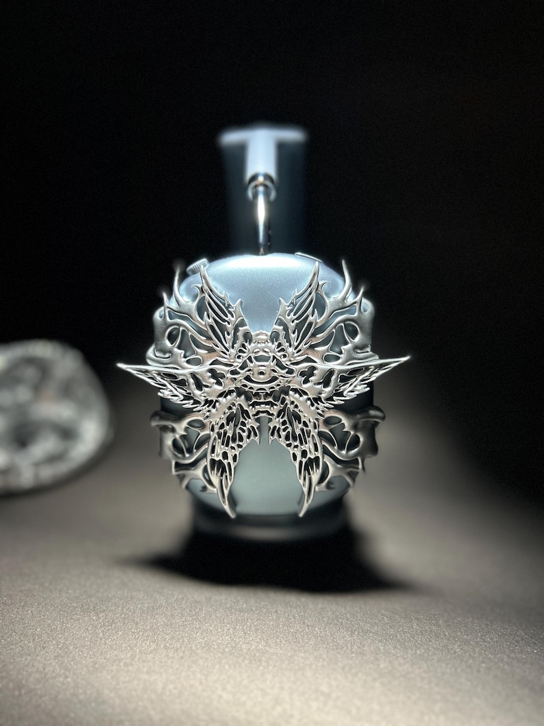 May include: A silver-colored headphone accessory with a detailed, intricate design resembling a butterfly. The accessory is attached to a light blue headphone.
