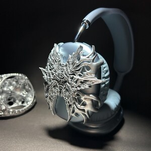May include: Silver headphones with a decorative, ornate, silver, winged design on the earcup. The headphones are light blue.