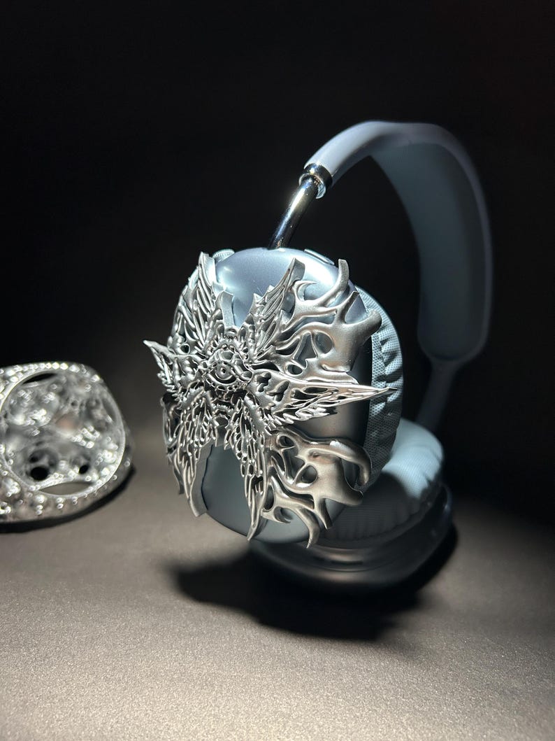 May include: Silver headphones with a decorative, ornate, silver, winged design on the earcup. The headphones are light blue.