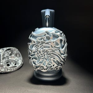 Può includere: Un paio di cuffie color argento con un design unico, intricato e astratto sul padiglione auricolare. Il design ricorda un motivo a spirale organico.