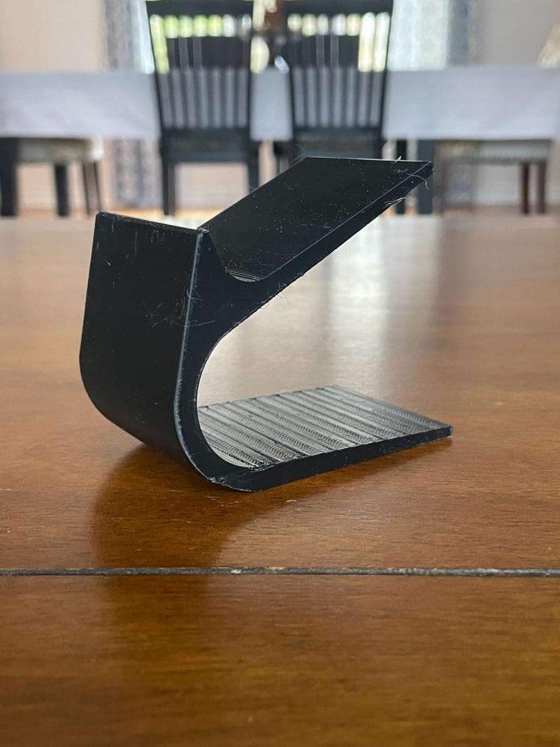 May include: A black, curved, 3D-printed object with a textured base. The object has a unique shape, with a raised, angled top and a curved, open design. The base features parallel lines, adding texture to the piece. The object is made of a matte black material.
