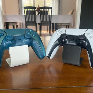 May include: Two game controllers, one teal and one white, are displayed on separate stands. The teal controller has a colour gradient, shifting from teal to blue. The white controller has black accents. Both controllers have black buttons, on a wooden surface.