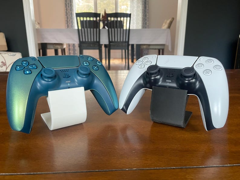 May include: Two game controllers, one teal and one white, are displayed on separate stands. The teal controller has a colour gradient, shifting from teal to blue. The white controller has black accents. Both controllers have black buttons, on a wooden surface.