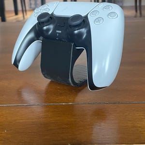 May include: A white and black PlayStation 5 DualSense wireless controller with a black wrist strap. The controller has black joysticks, buttons, and a touchpad. The controller is resting on a wooden surface.