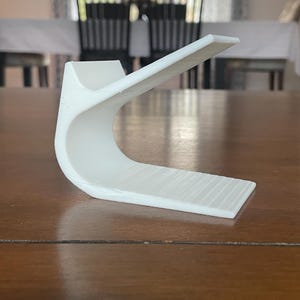 May include: A white, curved, 3D-printed object with a rectangular base and a raised, angled top. The object is made of a solid white material and sits on a wooden surface. The design is modern and minimalist.