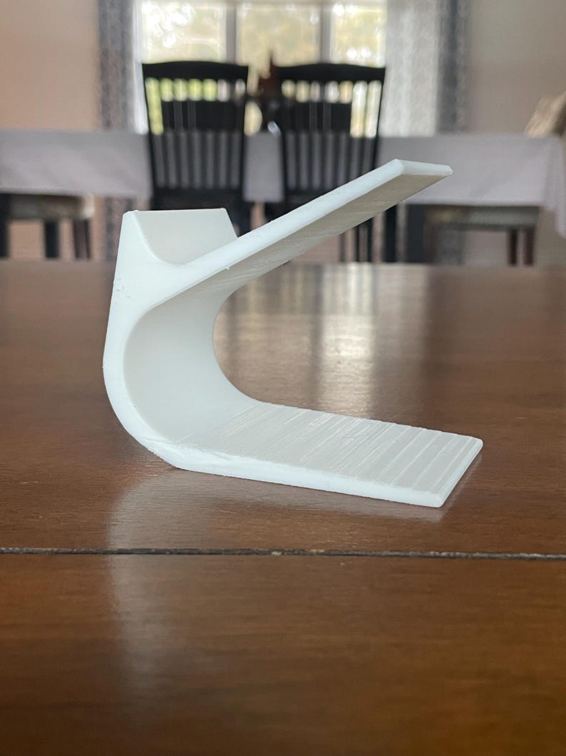 May include: A white, curved, 3D-printed object with a rectangular base and a raised, angled top. The object is made of a solid white material and sits on a wooden surface. The design is modern and minimalist.