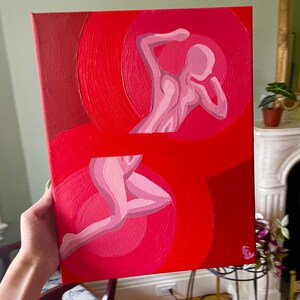May include: A red canvas painting featuring two stylized human figures in shades of pink. The figures are set against a backdrop of concentric red circles, creating a sense of movement and depth. The artwork is signed in the bottom right corner.