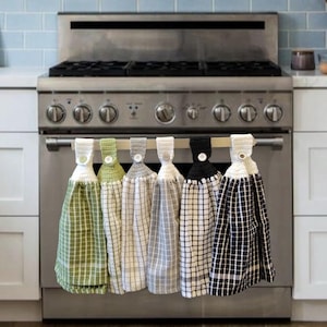 May include: Six kitchen towels with crocheted edges and button accents hanging on a rack above a stainless steel cooker. The towels are in various colours and patterns, including green, white, grey, and black.