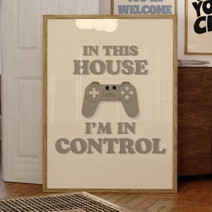 May include: A beige poster with a brown wooden frame. The poster has a gray video game controller with a smiling face on it. The text on the poster reads "In this house I'm in control."