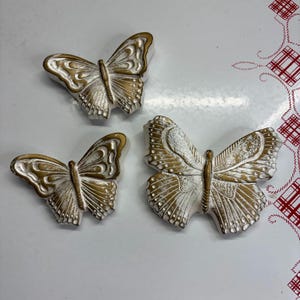 Op de afbeelding: Drie decoratieve vlinderbeeldjes in verschillende maten. Elke vlinder heeft een witte basis met goudkleurige accenten op de vleugels en het lichaam. De vlinders zijn gerangschikt op een wit oppervlak met een rood en wit patroon.