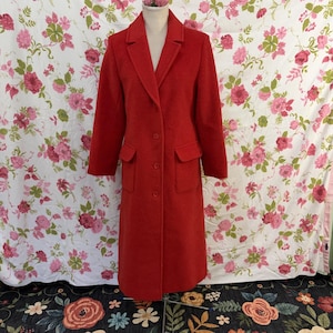 Peut inclure: Un long manteau rouge vif doté d'un col structuré, de manches longues et de trois boutons visibles sur le devant. Le manteau comprend deux poches à rabat rectangulaires sur les côtés et descend sous les genoux, dans un tissu uni et texturé.