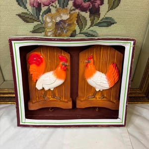 Può includere: Due statuine di gallo in legno da appendere al muro con vernice rossa, bianca e nera. I galli sono rivolti in direzioni opposte e sono montati su uno sfondo in legno marrone.