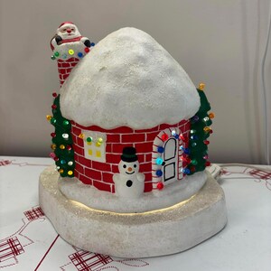 May include: A festive Christmas decoration featuring a red brick house with a snowy roof. A snowman and Santa Claus are visible. The house has a door and windows with colorful lights, and is surrounded by small Christmas trees.
