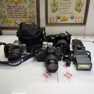 Peut inclure: Un ensemble d'équipement photographique comprenant un appareil photo Canon EOS argenté avec objectif et sangle, un appareil photo Minolta X-700 noir avec objectif, un boîtier Minolta noir, un sac photo Lowepro noir et un flash Minolta noir à tête rectangulaire.