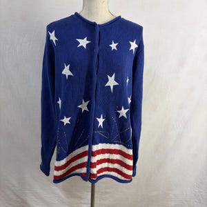 May include: A navy blue cardigan sweater with white star and red and white striped designs. The sweater has a button-up front and long sleeves. The bottom of the sweater features a red and white striped pattern, and the top has white stars.