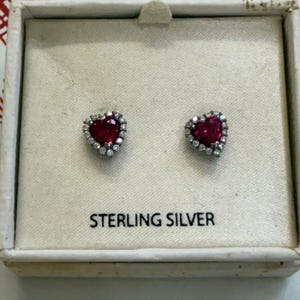 May include: A pair of heart-shaped stud earrings featuring a central red stone surrounded by a halo of small clear stones, set in sterling silver. The earrings are presented in a square box with the text STERLING SILVER printed on the base.