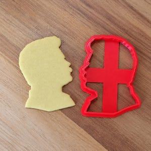 May include: A light yellow biscuit shaped like a human head in profile next to a red plastic biscuit cutter with the same silhouette, featuring a cross-shaped internal structure that creates a cutout design within the dough shape.
