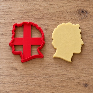 Puede incluir: Un cortador de galletas de plástico rojo con forma de perfil de cabeza humana y una estructura de soporte en cruz en su interior, junto a una pieza de masa de galleta de color beige claro con la misma forma sobre una superficie de madera.