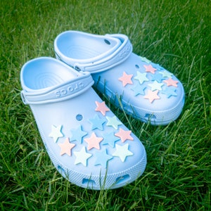 Op de afbeelding: Een paar lichtblauwe Crocs-klompen met een verscheidenheid aan pastelkleurige ster-charms eraan bevestigd. De klompen liggen op een bed van groen gras.