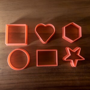 May include: Six different cookie cutters made of pink plastic. The cutters are shaped like a circle, square, heart, star, hexagon, and rectangle.