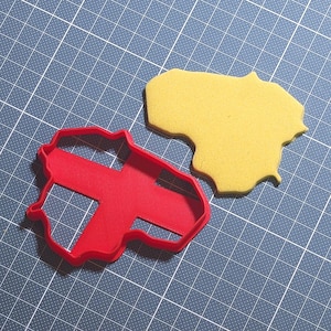 Pode incluir: Um cortador de biscoitos de plástico vermelho em forma de Lituânia, ao lado de um biscoito amarelo cortado na mesma forma. Os objetos estão em uma esteira de corte azul.