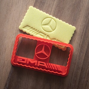 Könnte beinhalten: Ein roter Kunststoff-Ausstecher und ein gelber Keks mit dem AMG-Logo. Der Ausstecher ist rechteckig mit einem gewellten Rand und dem AMG-Logo. Der Keks hat die gleiche Form und das gleiche Logo, mit dem AMG-Logo geprägt.