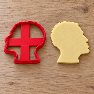 May include: A red plastic cookie cutter shaped like a human head in profile with a cross cutout, positioned next to a pale yellow cookie dough cutout of the same human head shape, showing the result of the tool on a flat surface.
