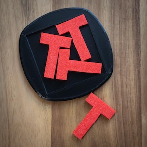 May include: A black tray with four red wooden puzzle pieces shaped like the letter T. One red T-shaped piece is outside the tray.