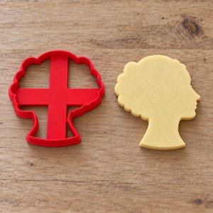 Può includere: Un tagliabiscotti in plastica rosso brillante a forma di profilo di testa umana con capelli ricci, posizionato accanto a una sagoma di pasta frolla giallo pallido della stessa forma, che mostra la silhouette di una persona con capelli strutturati.