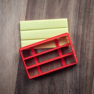 Può includere: Un tagliapasta di plastica rosso a forma di libreria con cinque ripiani. Il tagliapasta è sopra un rettangolo giallo chiaro.