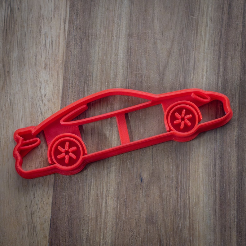 May include: Red plastic cookie cutter shaped like a car. The cutter has a detailed design with a front grill, headlights, and wheels.