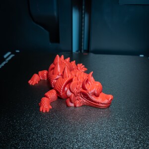 May include: A vibrant red 3D-printed dragon model. The dragon is depicted in a resting pose, with its limbs spread out. The model features intricate detailing, including scales and dorsal spikes. It rests on a black, textured surface.