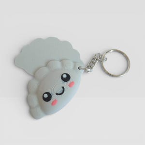 May include: A light gray dumpling-shaped keychain with a smiling face, black eyes, and pink cheeks. The keychain has a silver metal ring and chain. The dumpling has a ruffled edge and a textured surface.