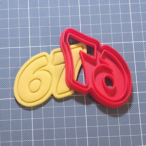 Op de afbeelding: Een gele koekjesvorm met de cijfers 6 en 7 naast een rode koekjesvorm met het cijfer 6. De koekjesvorm met de cijfers 6 en 7 is geel. De koekjesvorm met het cijfer 6 is rood. De koekjesvormen liggen op een blauwe snijmat.