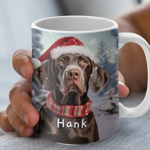 May include: White ceramic mug with a brown and white dog wearing a red Santa hat and a red and white scarf. The dog's name, "Hank", is written in white on the scarf.