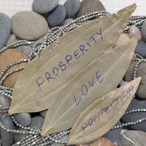 May include: Three dried bay leaves with handwritten words: "PROSPERITY", "LOVE", and "PROTECTION". The leaves rest on a bed of smooth stones and strands of silver beads. The background features various gray and tan stones.