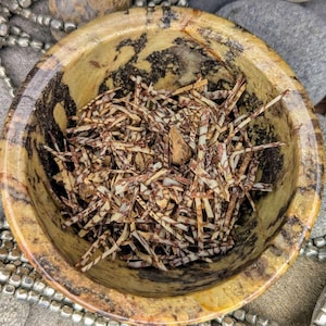 May include: A small, round, stone bowl with a mottled tan and black pattern, filled with dried, thin, brown and white pieces. The bowl is surrounded by grey stones and silver beads.