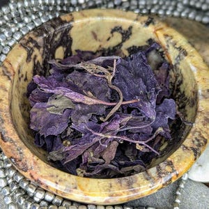 Può includere: Una piccola ciotola rotonda di pietra maculata piena di foglie viola secche. La ciotola ha un motivo marmorizzato marrone e beige. La ciotola è circondata da perline d'argento e pietre grigie lisce.