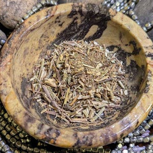 Op de afbeelding: Een close-up van een kleine, bruine kom gevuld met gedroogde kruiden en plantenmateriaal. De kom is gemaakt van een gemarmerd, lichtbruin steen met donkerbruine aderen.