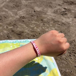 May include: A woven friendship bracelet featuring a repeating pattern of bright pink and white thread. The bracelet is constructed with a series of tight, interlocking knots that create a textured, wavy design around the circumference of the band.