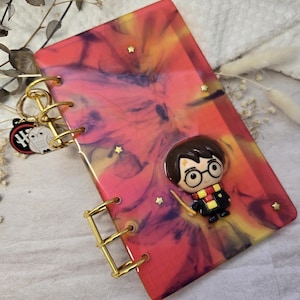 Peut inclure: Un carnet en résine fait à la main avec un motif tourbillonnant rouge, jaune et violet. Le carnet est orné d'une reliure dorée et d'un charm de personnage Harry Potter. De petites étoiles dorées ornent la couverture.