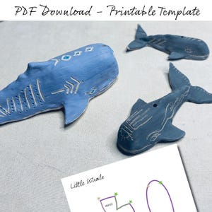 Könnte beinhalten: Drei blaue und graue Wal-Figuren mit weißen Markierungen. Die Wale sind aus Ton gefertigt und haben eine strukturierte Oberfläche. Der größte Wal hat einen blauen Körper mit weißen Markierungen und einen grauen Bauch. Die beiden kleineren Wale sind grau mit weißen Markierungen. Das Bild enthält auch eine druckbare Vorlage mit einer Strichzeichnung eines Wals und dem Wort "Kleiner Wal".