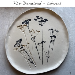 May include: A cream-colored ceramic plate with a raised rim, featuring a botanical design of black and tan wildflowers. The plate's surface has a textured appearance, and the text "PDF Download - Tutorial" is visible at the top.